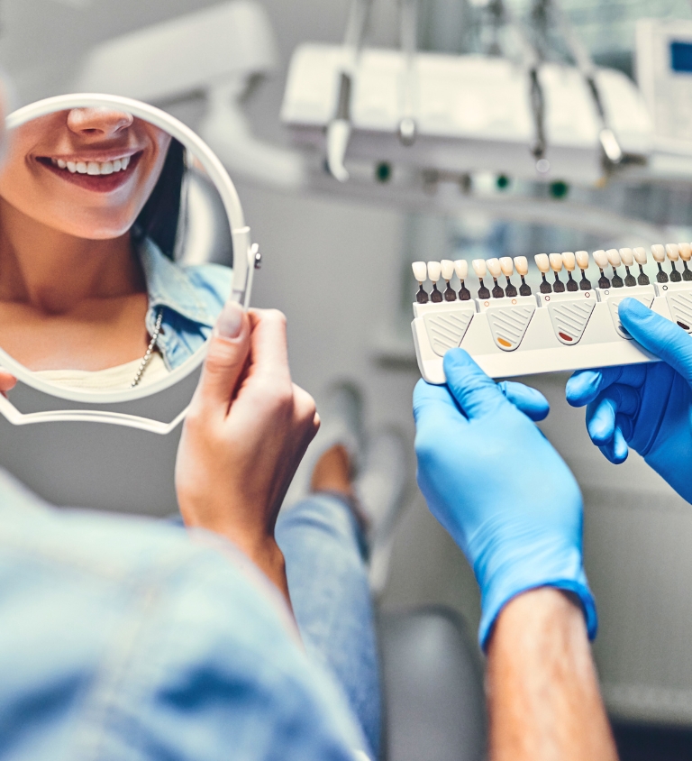 A dentist matching a patient's tooth shade while she holds a mirror in the dental chair